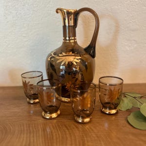 May include: A vintage brown glass decanter with a gold rim and floral design, accompanied by four matching shot glasses. The glasses and decanter have a gold rim. The set is displayed on a wooden surface.