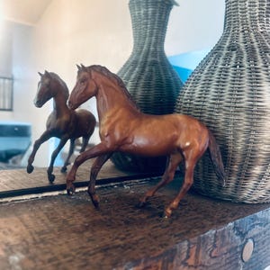 May include: A brown horse figurine with a raised front leg stands on a wooden surface, with its reflection visible in a mirror. A second horse figurine is in the background. Two grey wicker vases with greenery are also in the background.