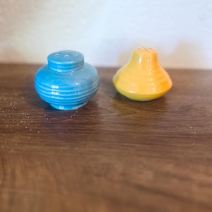 Puede incluir: Un par de saleros y pimenteros de cerámica. Uno es azul claro, de forma cilíndrica con estrías horizontales. El otro es amarillo, en forma de pera, y también tiene estrías horizontales. Ambos tienen pequeños agujeros en la parte superior.