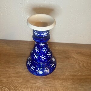 May include: A blue ceramic candle holder with a white top and snowflake design. The candle holder has a round base and a bulbous body. The snowflakes are white and evenly spaced around the body of the candle holder. The top is a shallow dish shape.