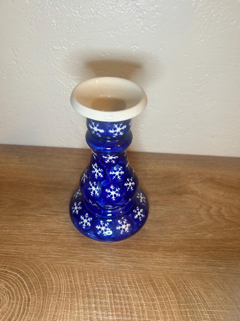 May include: A blue ceramic candle holder with a white top and snowflake design. The candle holder has a round base and a bulbous body. The snowflakes are white and evenly spaced around the body of the candle holder. The top is a shallow dish shape.