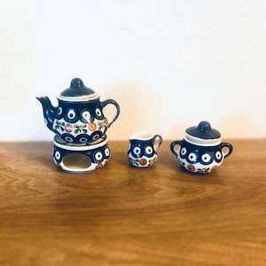 Op de afbeelding: Een miniatuur keramische theeset met een donkerblauwe basiskleur en witte en oranje bloempatronen. De set bevat een theepot met deksel, een melkkan, een suikerpot met deksel en een warmhoudstandaard. De set wordt gepresenteerd op een houten oppervlak.