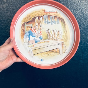 Puede incluir: Bandeja redonda de metal con borde rojo y una detallada ilustración de conejos en una escena interior acogedora. La ilustración muestra un conejo con chaqueta azul y otros conejos alrededor de una mesa. La bandeja tiene el texto "Tea & Tobacco".