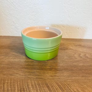 May include: A small, round, ceramic ramekin with a gradient green exterior and a light pink interior. The ramekin has a smooth, glossy finish and two horizontal ridges around the middle. It sits on a wooden surface.