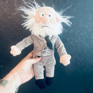 Può includere: Una bambola di peluche raffigurante un personaggio anziano con testa calva, capelli bianchi spettinati e una folta barba bianca. La bambola indossa una giacca da completo in velluto grigio con due bottoni, cravatta nera, pantaloni grigi e scarpe nere.