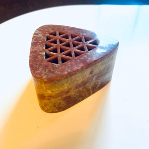 May include: A triangular, stone-like object with a geometric pattern of cut-out triangles on the top surface. The object has a reddish-brown color with lighter brown and yellow bands. The sides are smooth and the overall shape is a truncated triangle.