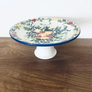 May include: A ceramic pedestal plate featuring a cream-colored surface with a hand-painted floral pattern in shades of red, yellow, green, and blue. The circular plate has a blue rim and is supported by a solid white, flared cylindrical base.