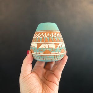 May include: A small, hand-painted ceramic vase with a turquoise top and a patterned design in orange, white, and turquoise. The vase has a rounded shape and is held in a hand against a dark background. The design features geometric shapes and lines.