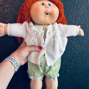 May include: A doll with bright orange yarn hair and blue eyes, wearing a white knit cardigan with green and pink embroidery details over a green and white gingham romper featuring white lace trim at the leg openings and a small fabric tag.