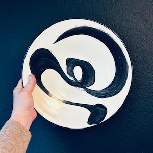 Puede incluir: Un plato de cerámica blanco con un diseño abstracto negro. El diseño es una serie de líneas y curvas en espiral.