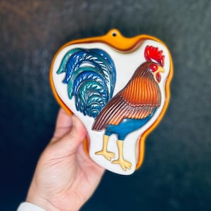 May include: A ceramic rooster wall hanging with a heart-shaped design. The rooster features a red comb, orange and brown feathers, blue legs, and a blue and green tail. The border is orange.