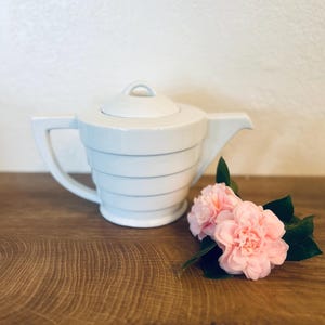 May include: A white ceramic teapot with a lid, featuring horizontal gray lines. The teapot has a handle and a spout. Pink flowers with green leaves are placed next to the teapot on a wooden surface.