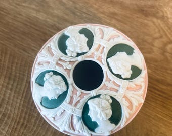 Puede incluir: Una pieza decorativa redonda de cerámica con un agujero negro central y cuatro óvalos verdes, cada uno con un perfil de camafeo blanco. Tiene un borde rosa con detalles florales en relieve blanco.