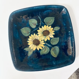 Op de afbeelding: Een vierkant keramieken bord met een diep, glanzend marineblauw glazuur. Het midden is voorzien van een handgeschilderd ontwerp van twee gele zonnebloemen met bruine harten, omringd door diverse groene bladeren met zichtbare donkere nerven.