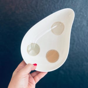 May include: A teardrop-shaped white ceramic dish featuring three distinct circular accents in muted shades of beige, light gray, and tan. The bowl has a smooth, glossy finish and a tapered point at one end, providing a simple and functional design.