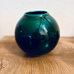 以下が含まれることがあります： 光沢のあるエメラルドグリーンの花瓶。球形をしており、広い開口部があります。木製の表面に置かれています。花瓶の反射面は周囲の環境を映し出します。インテリア装飾に適した装飾品です。