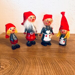 May include: Four wooden gnome figurines with red hats, black coats, and red or black shoes. One gnome has a white beard and carries a lantern. Another wears glasses and an apron with a heart design. The figurines are displayed on a wooden surface.