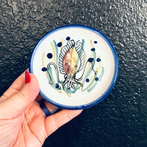 May include: A small, round ceramic dish with a hand-painted squid design. The dish has a white interior with a blue rim and a blue squid illustration with brown, yellow, and green accents. The dish is held in a hand.