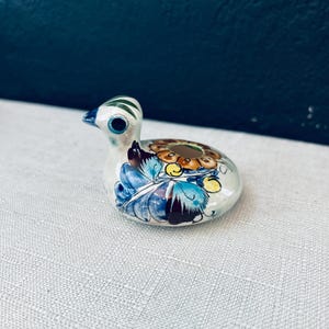 Puede incluir: Figura de pato de cerámica con base blanca, con diseños florales azules, marrones y amarillos. El pato tiene pico y ojo azules, con rayas verdes en la cabeza. Un artículo decorativo.