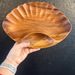 May include: A wooden serving dish with a scalloped edge and a natural wood grain finish. The dish has a shallow, oval-shaped indentation on one side. The dish is designed for serving appetizers or snacks.