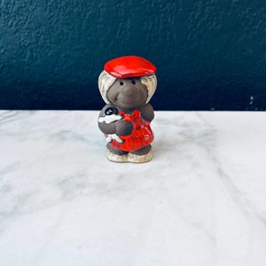 May include: A small ceramic figurine of a person wearing a red hat and a red and white patterned dress. The figurine is holding a small black and white dog.