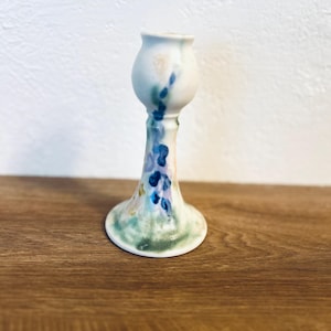 Può includere: Un portacandele in ceramica bianca con base svasata e parte superiore bulbosa. La superficie presenta un design dipinto a mano in stile acquerello con striature verticali di pigmenti blu, verdi e giallo tenue che si fondono nella finitura bianca.