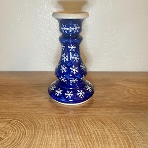 May include: A blue ceramic candlestick with white snowflake designs. The candlestick has a wide base, a narrow neck, and a flared top. The background is a light-colored wall and a wooden surface.