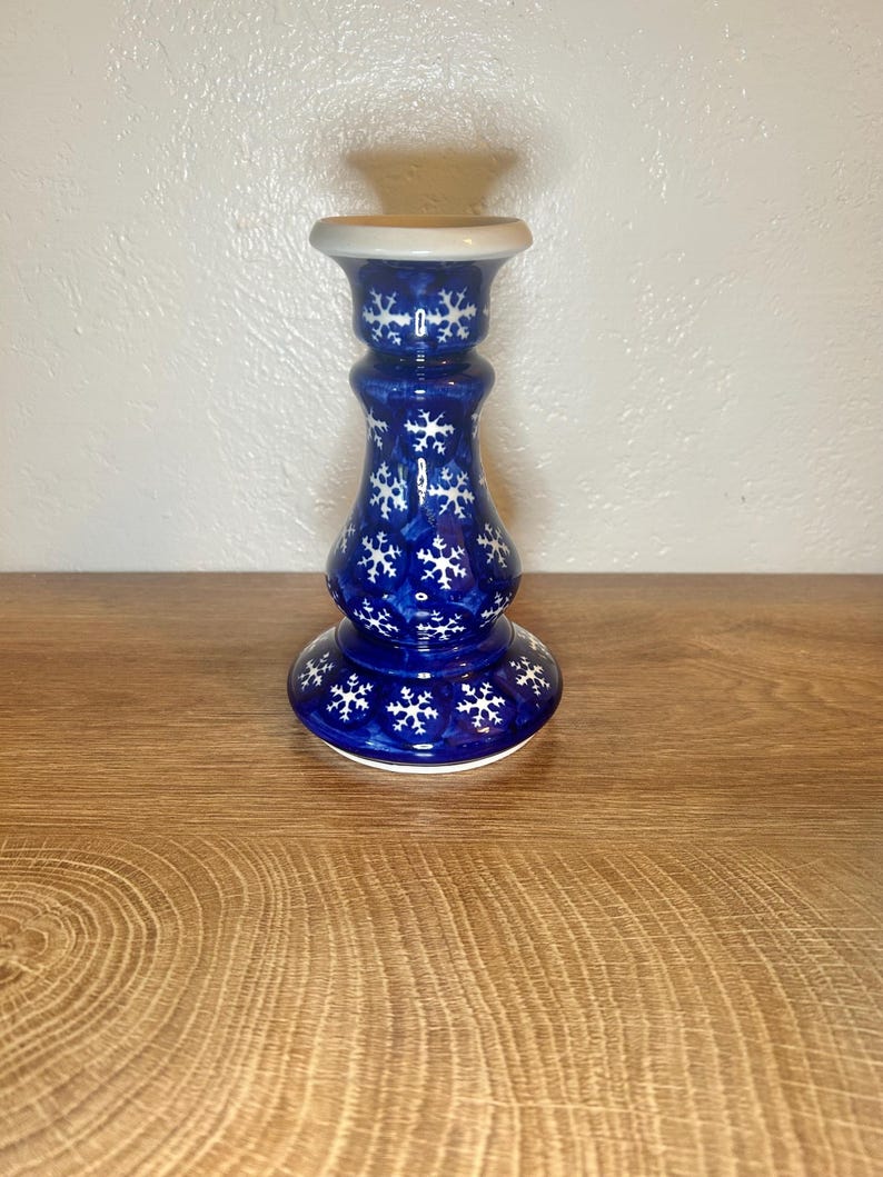 May include: A blue ceramic candlestick with white snowflake designs. The candlestick has a wide base, a narrow neck, and a flared top. The background is a light-colored wall and a wooden surface.