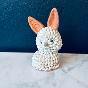 May include: A white ceramic bunny figurine made of small seashells with orange ears. The bunny is sitting on a white marble surface.