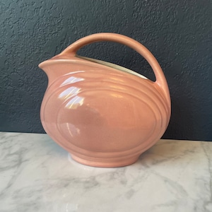 Puede incluir: Una jarra de cerámica rosa con cuerpo redondeado y asa curva. La jarra tiene un acabado brillante y un sutil patrón de nervaduras.