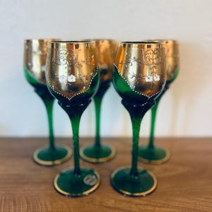 May include: Set of five green wine glasses with gold rims and ornate gold detailing on the bowls. The bowls feature white floral accents and are set on green stems and bases. A decorative glassware set.