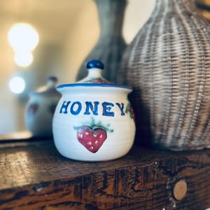 May include: A white ceramic honey pot with a blue lid and the word "HONEY" in blue lettering. A red strawberry is painted below the word. The pot is on a wooden shelf.