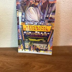 Puede incluir: Un libro de cocina titulado "The Stinking Cookbook" con una colorida ilustración de un bulbo de ajo y un paisaje urbano. La portada presenta el texto "The Stinking Rose" y un diseño artístico vibrante.