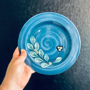 Puede incluir: Un plato de cerámica azul pintado a mano con un diseño floral. El plato presenta un patrón en espiral y una rama pintada con hojas blancas y pequeños acentos verdes. También hay una flor en blanco y negro pintada en el plato.