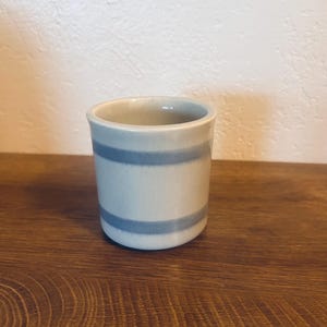 Pode incluir: Uma pequena chávena cilíndrica de cerâmica em azul claro e branco. A chávena apresenta duas riscas azuis horizontais no meio. Está colocada numa superfície de madeira.