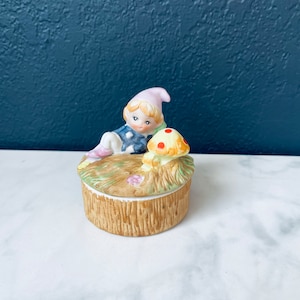 Puede incluir: Una caja de cerámica para guardar pequeños objetos con una pequeña figura de elfo sentada junto a un hongo. El elfo lleva un atuendo azul y blanco con un sombrero rosa. El hongo es amarillo con manchas rojas. La caja está decorada con hierba verde y flores.