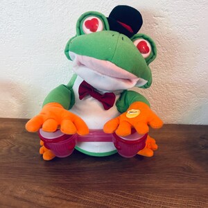 May include: A plush green frog toy wearing a black top hat and a red bow tie. The frog has heart-shaped red pupils in its eyes and orange hands resting on a pair of small, translucent pink bongo drums. A yellow circular sticker is on one hand.
