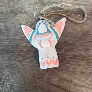 May include: A white ceramic angel ornament with painted coral, teal, and blue details. The angel has outstretched wings, hands pressed together in prayer, and a jute twine loop attached to the top for hanging purposes.