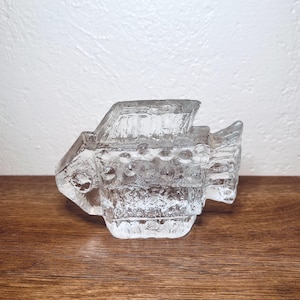 May include: A clear, textured glass fish sculpture. The fish has a rectangular body with a textured surface and fins. The sculpture is sitting on a wooden surface.