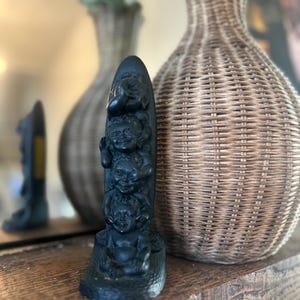 May include: A black sculpture of four whimsical faces stacked vertically. The sculpture is on a wooden surface, with a woven vase and a mirror in the background. The sculpture is a unique decorative item.
