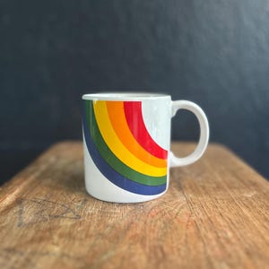 Pode incluir: Caneca de cerâmica branca com um design de arco-íris colorido. O arco-íris se estende pela lateral da caneca, exibindo faixas de vermelho, laranja, amarelo, verde e azul. A caneca tem uma alça curva e está sobre uma superfície de madeira.