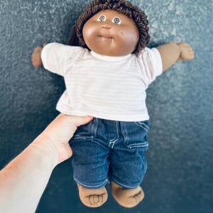 May include: A doll with a round face, dark brown skin, and short dark brown yarn hair. The doll wears a white short-sleeved shirt with thin horizontal stripes and dark blue denim trousers with visible white stitching on the pockets and legs.