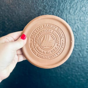 May include: A round, terracotta-colored Longaberger Pottery Button Basket Brick. The surface features embossed text reading Button Basket Brick, Longaberger Pottery, Made in the U.S.A., and Use With Extreme Care When Hot, surrounding a central logo.