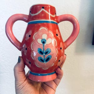 May include: A hand-painted ceramic vase with two handles. The vase is primarily red with pink and blue accents. It features a floral design in pink, white, and blue. The vase is held in a hand.