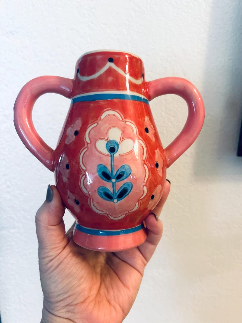 May include: A hand-painted ceramic vase with two handles. The vase is primarily red with pink and blue accents. It features a floral design in pink, white, and blue. The vase is held in a hand.
