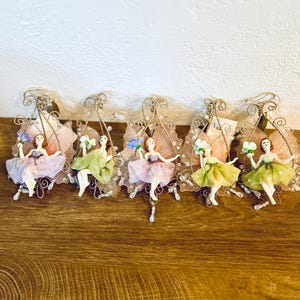 May include: A collection of five fairy ornaments. Each fairy is dressed in a pastel-colored dress and holds a small bouquet of flowers. The fairies are suspended from a decorative gold-colored wire frame with beaded accents and pink wings.