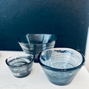 May include: Three hand-blown glass bowls in varying sizes. The bowls are a smoky gray color with a marbled effect. The bowls are displayed on a white surface against a dark blue background. The bowls have a simple, elegant design.