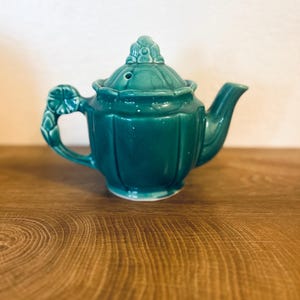 May include: A teal ceramic teapot with a matching lid. The teapot has a curved spout, a handle with a leaf design, and vertical ridges along the body. The lid is topped with a decorative floral element. The teapot is on a wooden surface.