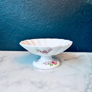 Può includere: Un piatto da portata bianco con piedistallo e bordo smerlato. Il piatto presenta un motivo floreale con rose rosa e verdi sulla base e sul bordo. Il piatto è posto su uno sfondo blu scuro e una superficie di marmo.