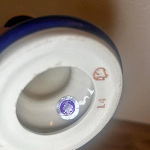 May include: A ceramic vase with a white base and a dark blue rim. The bottom of the vase has a sticker with text and a number 14 in brown ink. The vase is handcrafted.